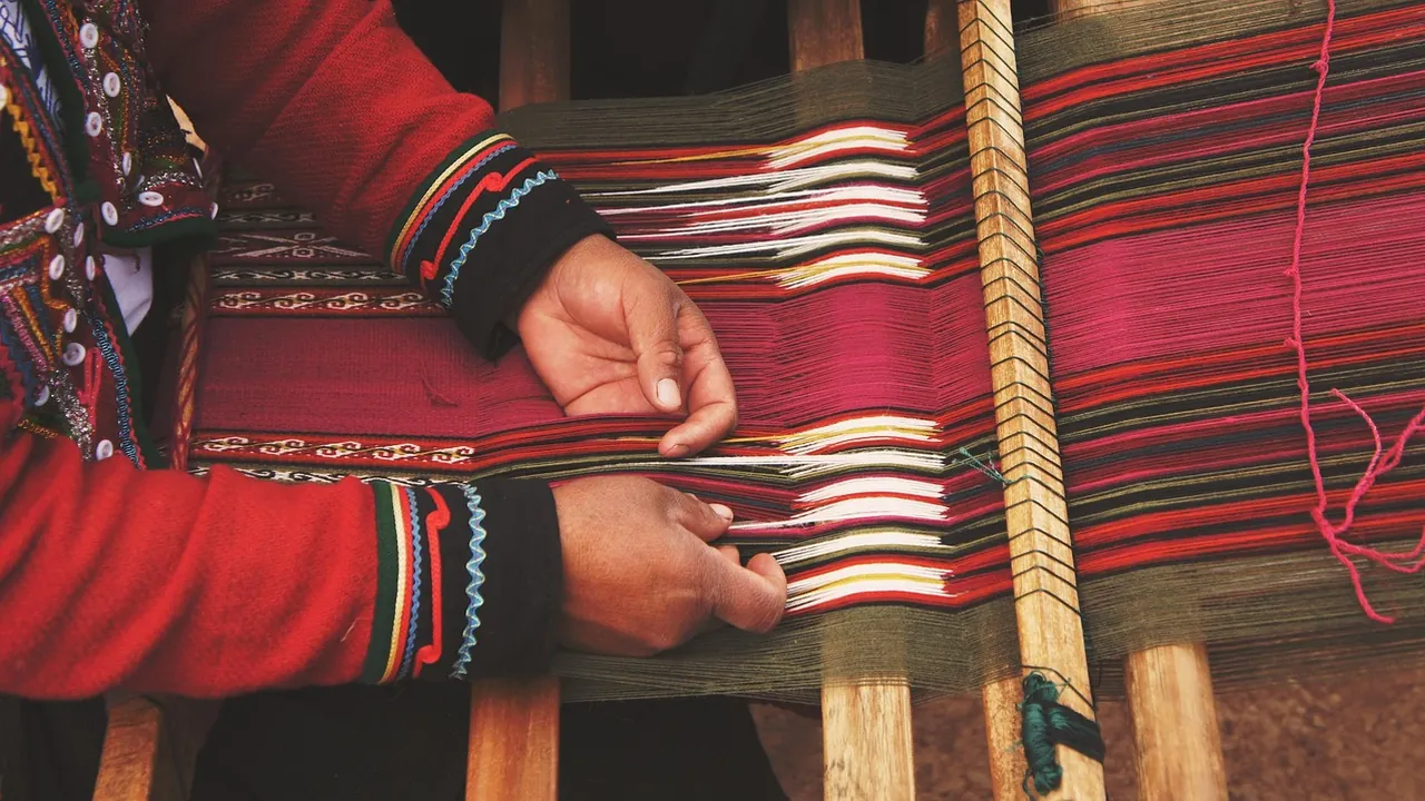 Des mains tissent une étoffe rouge à motifs traditionnels sur un métier en bois